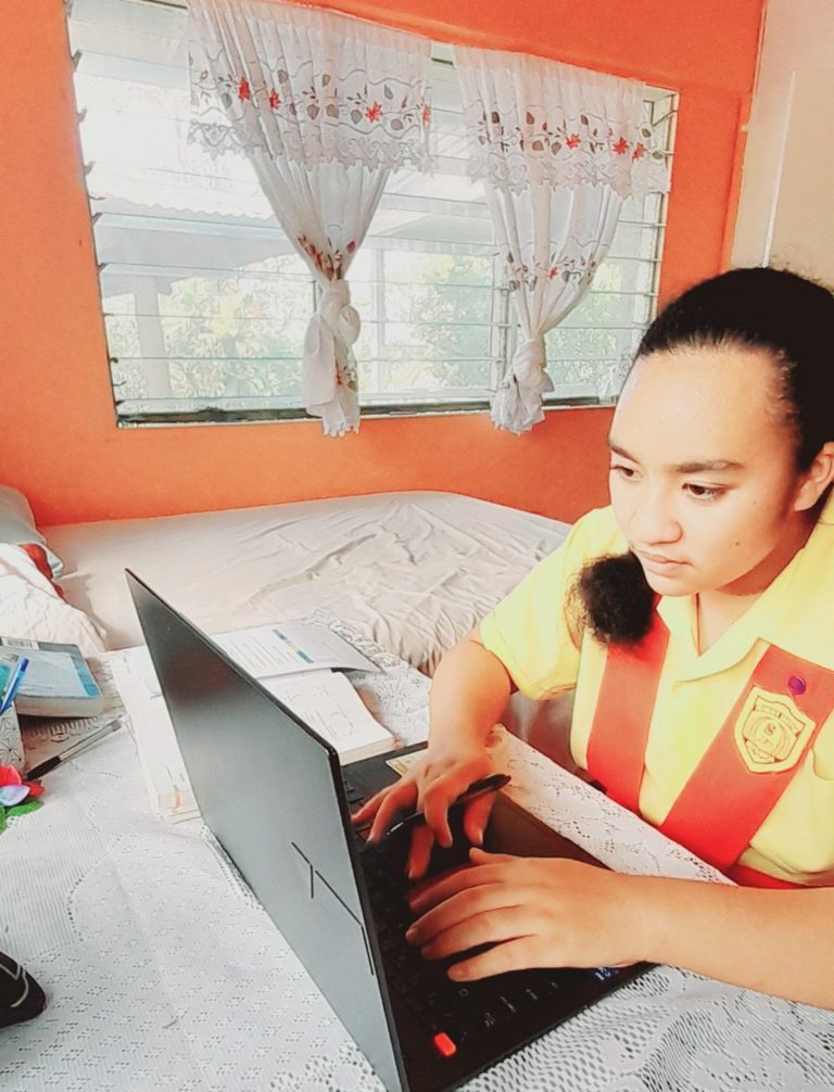 A student in Samoa sits at her bedroom desk, dressed in school uniform, enjoying studying on her laptop sponsored by Dauntless Blue.
